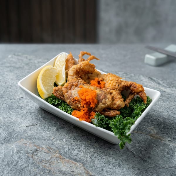 Crispy fried soft-shell crab garnished with orange roe, lemon wedges, and curly lettuce in a white rectangular dish.