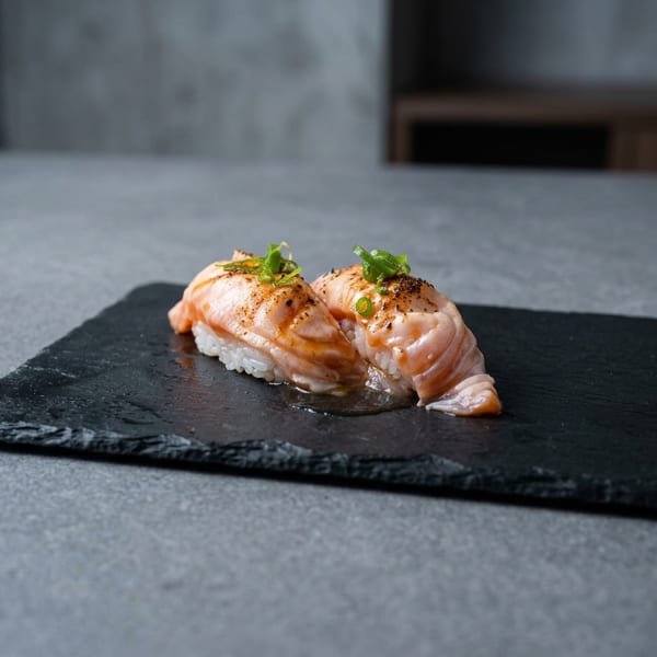 Two seared salmon sushi pieces topped with green onions on a black slate serving board.