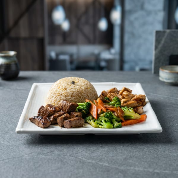 Square plate featuring a mound of rice, grilled meat, diced tofu, and colorful broccoli and carrots on a restaurant table.