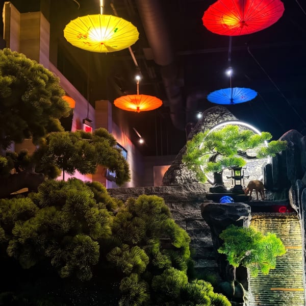 Japanese-inspired restaurant interior with sculpted pine trees, illuminated umbrellas, rock landscaping, and a cascading water feature.