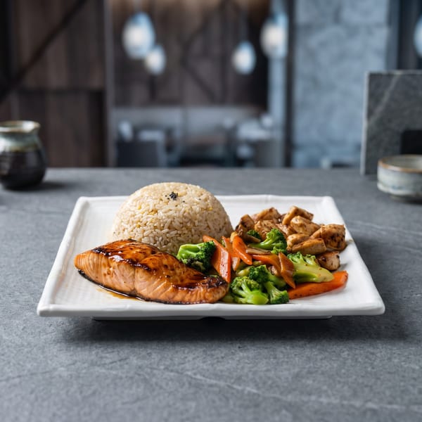 White plate featuring glazed salmon, fried rice, grilled chicken, and colorful steamed vegetables on a restaurant table.