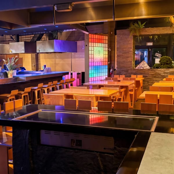 Modern restaurant interior with sushi bar, colorful lighting, wooden tables, bar stools, and a large central dining counter.