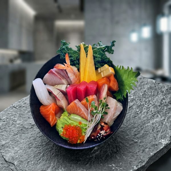 Colorful sashimi bowl with various fish, shrimp, avocado, roe, and greens on a stone surface.