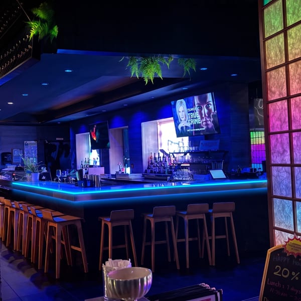 Restaurant bar with vibrant blue neon lighting, bar stools, bottles, and TVs.