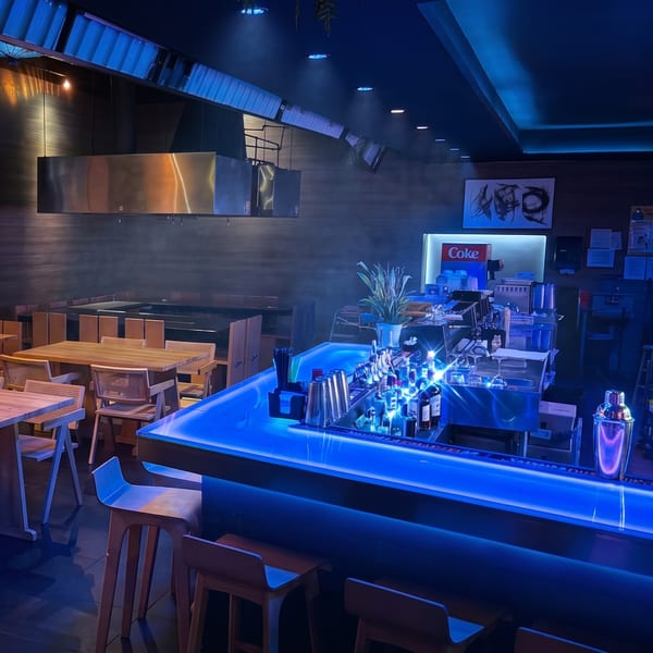 Modern restaurant interior with a glowing blue bar, bar stools, and dining tables in a smoky atmosphere.