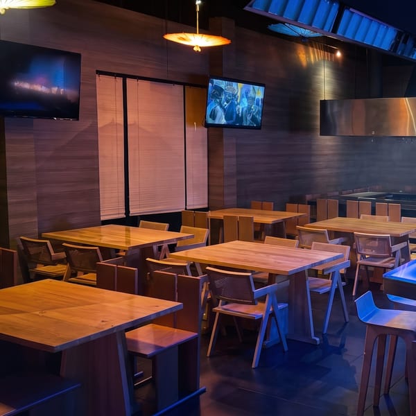 Dimly lit restaurant dining area with light wooden tables, chairs, and wall-mounted screens under decorative lamps.