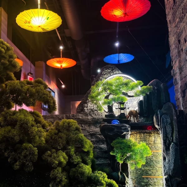 Restaurant interior with colorful hanging umbrellas, a lighted waterfall, bonsai tree, and deer statue.