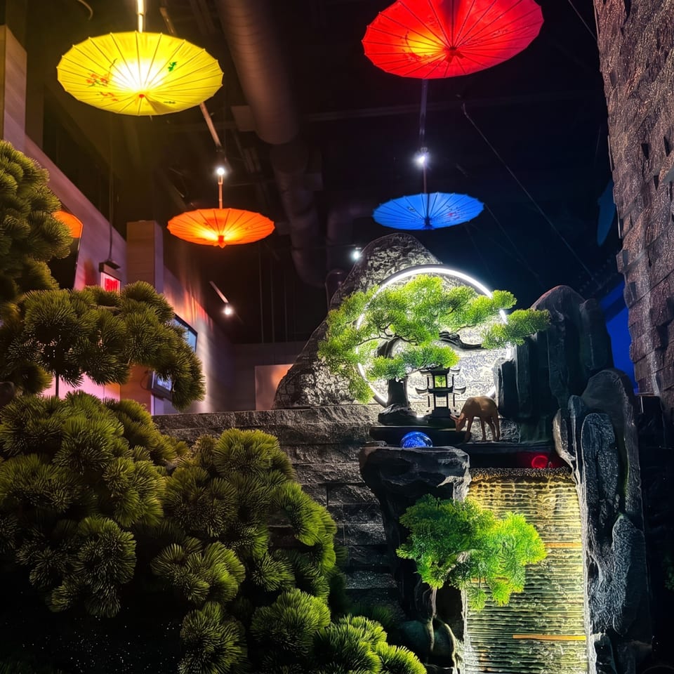 Restaurant interior with colorful hanging umbrellas, a lighted waterfall, bonsai tree, and deer statue.